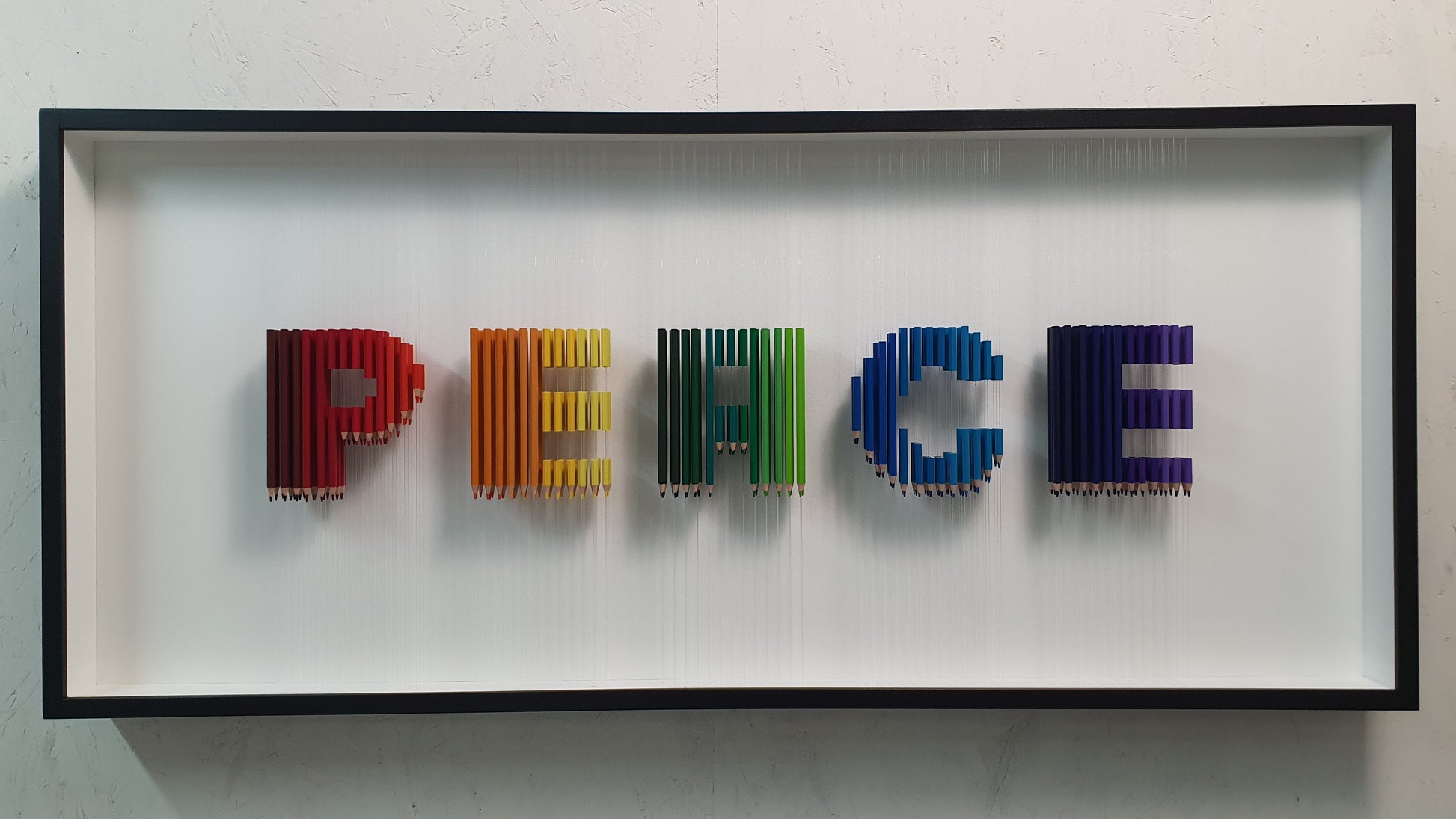 Arcobaleno Sospeso conceptual artwork – suspended rainbow-colored pencils forming the word PEACE, front view