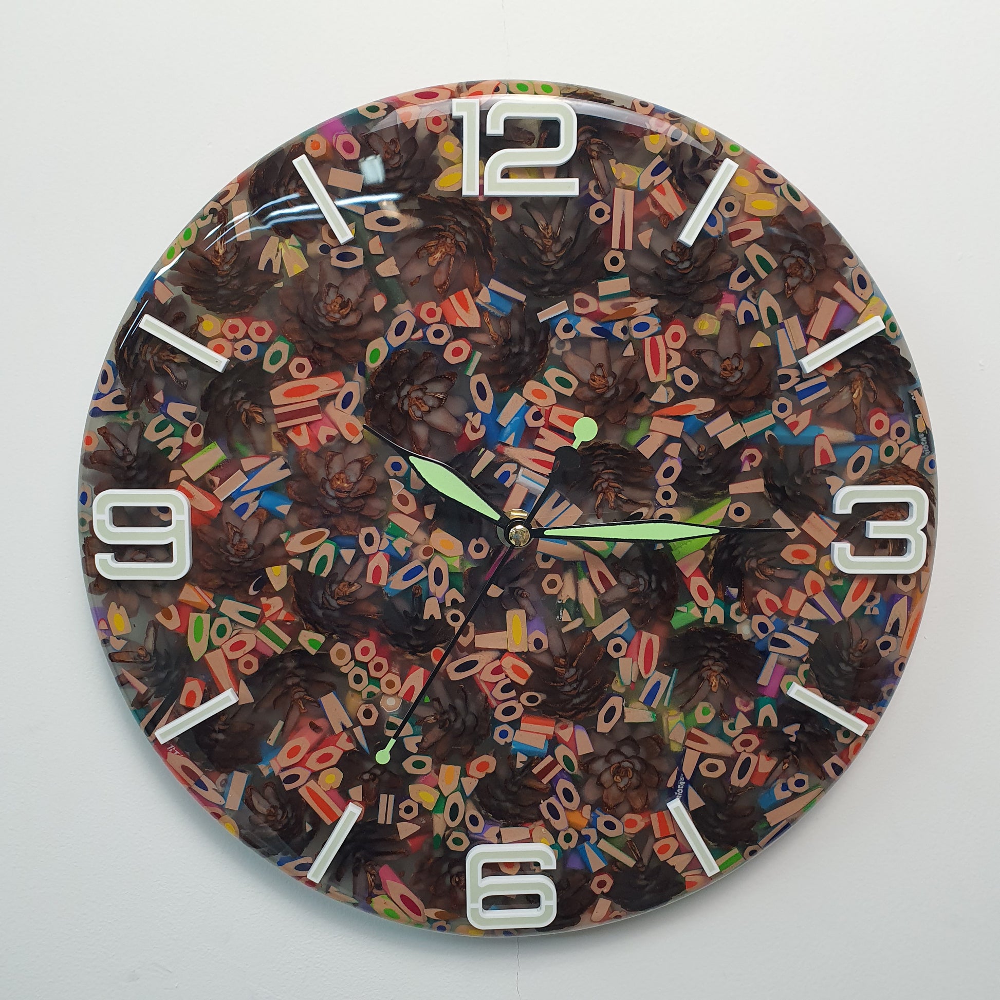Front view of the “Pine & Color Fusion” wall clock made from colorful pencils and pinecones embedded in epoxy resin, with glowing white plexiglass numbers.