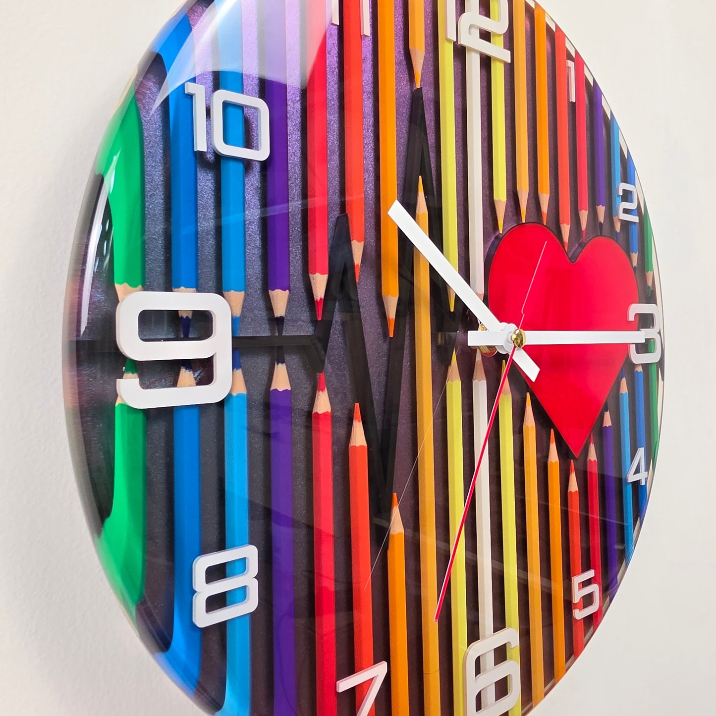 Modern artistic wall clock made with real colored pencils embedded in resin, white acrylic numbers, and a heartbeat design symbolizing imagination and creativity.